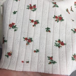May include: Close-up of a cream-colored ribbed fabric with a floral pattern. The pattern features small red roses with green leaves scattered across the surface. The fabric has a textured, vertical ribbed design.