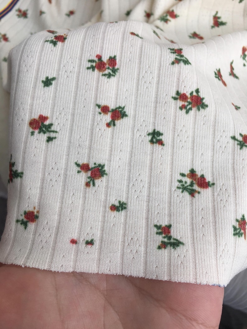 May include: Close-up of a cream-colored ribbed fabric with a floral pattern. The pattern features small red roses with green leaves scattered across the surface. The fabric has a textured, vertical ribbed design.