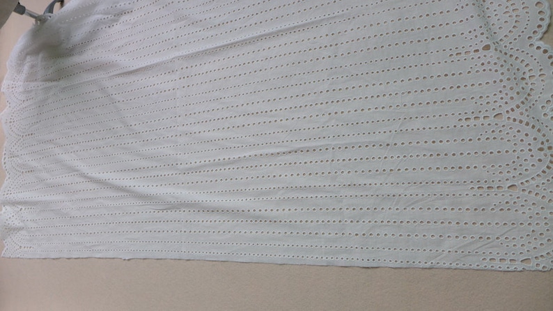 May include: White cotton fabric with a delicate eyelet pattern. The fabric has vertical rows of small holes, creating a lacy effect. The edges of the fabric are scalloped.