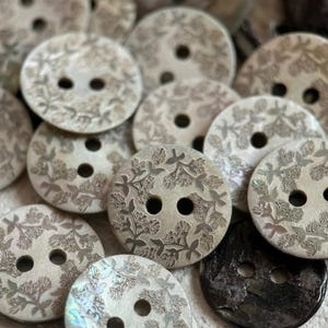 May include: A collection of round, two-hole buttons featuring an engraved floral pattern. Most buttons are a light, pearlescent off-white color with detailed botanical etchings, while one button is a solid, dark, glossy black material.