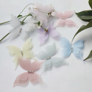 May include: A collection of delicate, pastel-colored butterfly embellishments. Each butterfly features layered, sheer wings in shades of white, pink, yellow, blue, and lavender, with a sparkling rhinestone center. Ideal for craft projects or adding a touch of whimsy.