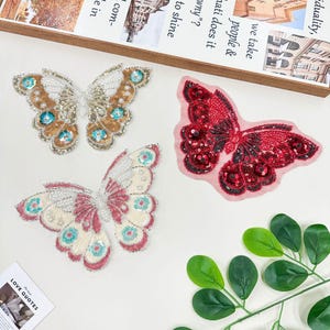 Può includere: Tre applicazioni decorative a farfalla. Una è oro e bianca con accenti turchesi, un'altra è rossa con dettagli neri e la terza è bianca e rosa con accenti turchesi. Le applicazioni sono impreziosite da perline e paillettes. Un rametto di foglie verdi è nell'angolo.