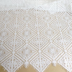 Könnte beinhalten: Weißer Spitzenstoff mit einem sich wiederholenden geometrischen Muster aus Quadraten und floralen Designs. Die Spitze hat einen gewellten Rand.