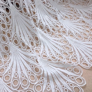 May include: White lace fabric with a peacock feather design. The fabric features a repeating pattern of open circles and delicate lines, creating a textured and airy appearance. The lace is likely used for apparel or home decor.