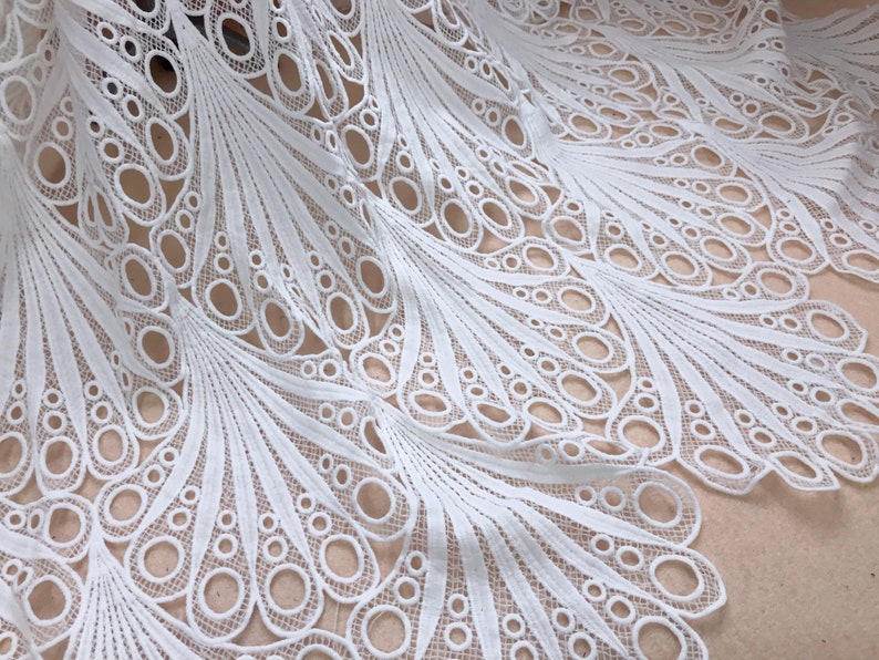 May include: White lace fabric with a peacock feather design. The fabric features a repeating pattern of open circles and delicate lines, creating a textured and airy appearance. The lace is likely used for apparel or home decor.