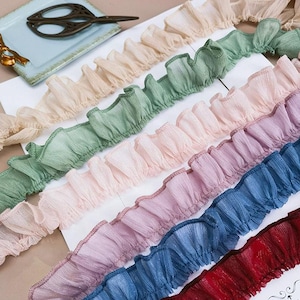 Könnte beinhalten: Verschiedene Rüschenborten in Beige, Grün, Rosa, Lila, Blau und Rot. Die Borten sind in parallelen Reihen angeordnet, um die Textur und Farbvariationen zu zeigen. Eine Schere und eine kleine blaue Schale sind im Hintergrund zu sehen.