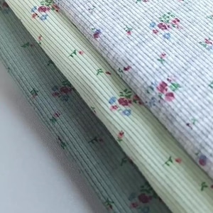 Pu&ograve; includere: Primo piano di tre campioni di tessuto con una trama a coste e un motivo floreale. I tessuti sono in tonalit&agrave; di grigio, verde e blu, con piccoli fiori rosa, blu e verdi sparsi sulla superficie. Il tessuto &egrave; ripiegato su se stesso.