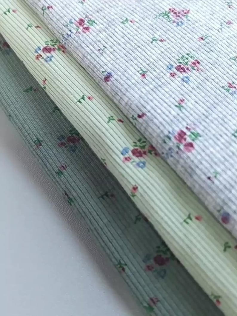 Pu&ograve; includere: Primo piano di tre campioni di tessuto con una trama a coste e un motivo floreale. I tessuti sono in tonalit&agrave; di grigio, verde e blu, con piccoli fiori rosa, blu e verdi sparsi sulla superficie. Il tessuto &egrave; ripiegato su se stesso.