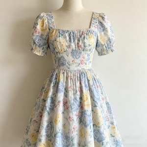 May include: A floral print dress with a square neckline and short, puffed sleeves. The dress features a fitted bodice and a full skirt. The fabric is white with blue, yellow, and pink rose patterns. The dress is displayed on a mannequin.
