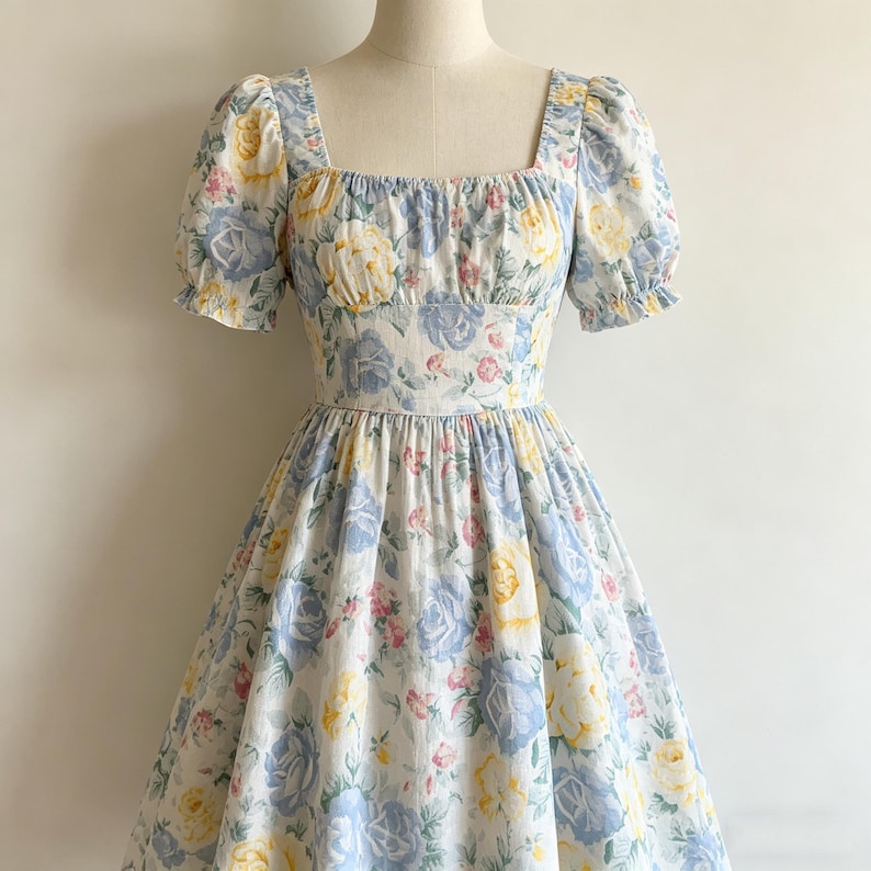 May include: A floral print dress with a square neckline and short, puffed sleeves. The dress features a fitted bodice and a full skirt. The fabric is white with blue, yellow, and pink rose patterns. The dress is displayed on a mannequin.