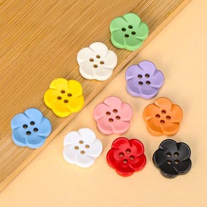 Könnte beinhalten: Eine Sammlung von zehn blumenförmigen Knöpfen in verschiedenen Farben, darunter Hellgrün, Weiß, Gelb, Hellblau, Lila, Rosa, Orange, Rot und Schwarz. Jeder Knopf verfügt über vier mittige Löcher, die in einem quadratischen Muster angeordnet sind.