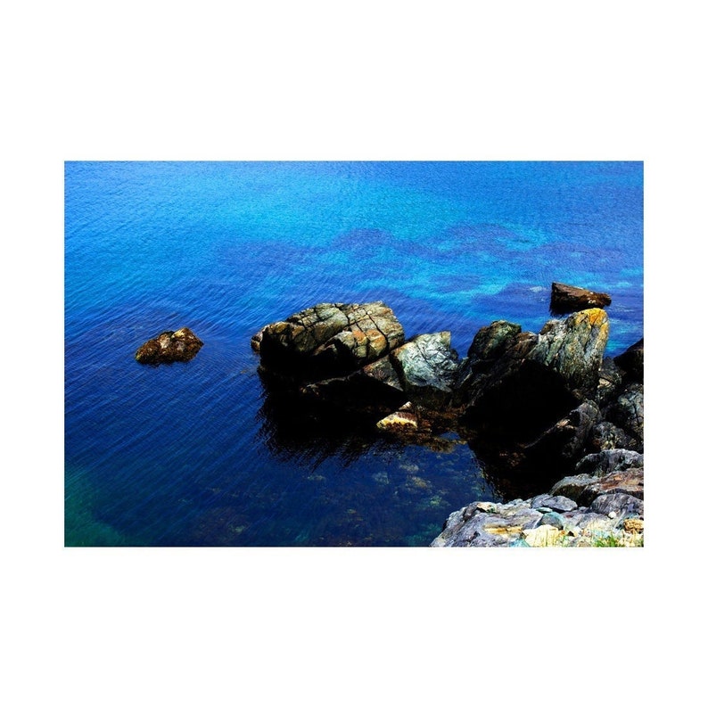 K&ouml;nnte beinhalten: Ein felsiger Ausl&auml;ufer ragt in ein klares blaues Gew&auml;sser. Die Felsen sind eine Mischung aus Braun, Grau und Gr&uuml;n, und das Wasser ist tiefblau.