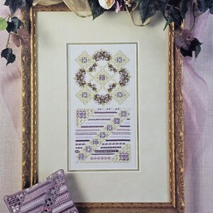 May include: A framed cross-stitch pattern with a floral design in shades of lavender, cream, and brown. The frame is gold-colored, and the image includes a matching lavender-colored needlepoint pillow. The text "I Will Wear Lavender" is visible.