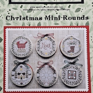 May include: A product image featuring six Christmas Mini-Round cross-stitch designs. Each round has a different holiday-themed design, including a bird, the word "Noel", a boot, a house, and a snowflake. The background is a light green with pine needles.