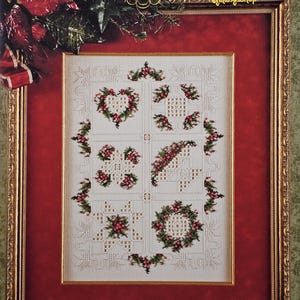 May include: A framed cross-stitch pattern with a Christmas theme. The design features various holiday motifs, including a heart, cross, and wreath, all adorned with red berries and green foliage. The frame is gold, set against a red background.