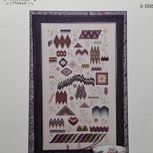 May include: A framed "Bargello Sampler" cross-stitch pattern with various geometric designs in shades of red, purple, and blue. The text "BARGELLO SAMPLER" is at the top. The frame has a dark, ornate pattern. A teacup with floral designs is in the lower right corner.
