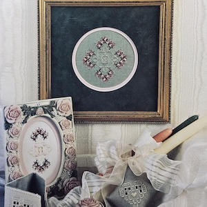 May include: An image featuring cross-stitch projects and supplies. A framed piece with a circular design is centered. Other items include a rose-adorned frame, a small box, and a decorative pillow, all with cross-stitch details. The words "CROSS 'N PATCH" are visible.