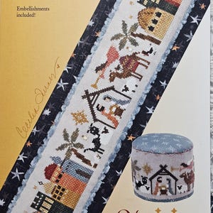 May include: A cross-stitch pattern featuring a Nativity scene with buildings, palm trees, and animals. The design includes the text "Nativity tiny town" and the Heart in Hand Needleart logo. Embellishments are included.
