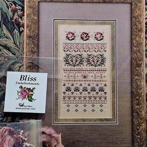 May include: A framed cross-stitch sampler titled Bliss featuring floral motifs, geometric borders, and text. Includes a small plastic bag labeled Bliss Embellishments containing pink embroidery thread, purple beads, and white pearls for needlework.