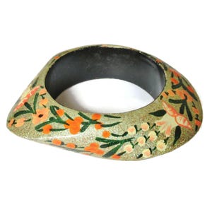 May include: A teardrop-shaped bangle bracelet with a textured, glittery gold finish. The surface is decorated with hand-painted orange flowers, green leaves, and white floral buds. The interior of the bangle is a solid, smooth matte black.