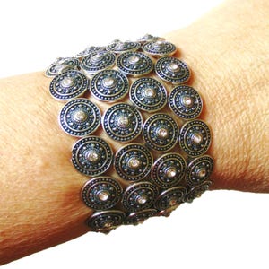 May include: A wide cuff bracelet composed of multiple rows of circular, silver-toned metal discs. Each disc features a textured, dark-oxidized finish with a raised center point and a small, clear, sparkling rhinestone embedded in the middle.