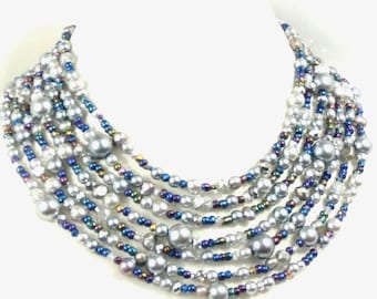 May include: A multi-strand necklace featuring strands of gray pearls and smaller beads in shades of blue, silver, and iridescent colors. The necklace has a layered design, creating a full and elegant look. The pearls are of varying sizes.