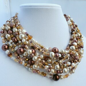 May include: A multi-strand statement necklace featuring a dense arrangement of round beads in shades of cream, champagne, bronze, and dark brown. The design incorporates a mix of smooth pearls, metallic-finish spheres, and small faceted crystals.