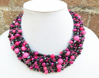 May include: A multi-strand statement necklace featuring a dense arrangement of beads in varying sizes and textures. The design incorporates matte and glossy hot pink beads, faceted black crystals, and smooth metallic charcoal grey spherical beads.