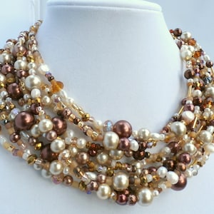 May include: A multi-strand statement necklace featuring a dense arrangement of round beads in shades of cream, champagne, and dark chocolate brown. The design incorporates a mix of smooth faux pearls, faceted crystal beads, and small glass seed beads.