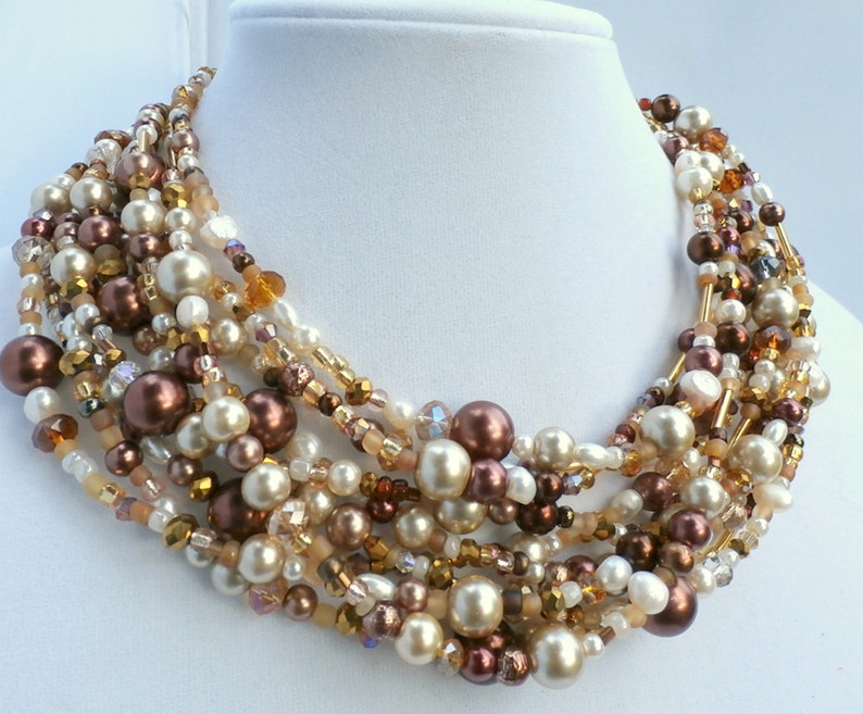 May include: A multi-strand statement necklace featuring a dense arrangement of round beads in shades of cream, champagne, and dark chocolate brown. The design incorporates a mix of smooth faux pearls, faceted crystal beads, and small glass seed beads.