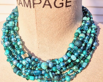 May include: Multi-strand turquoise and teal beaded necklace with various sizes and shapes of beads. The necklace is layered and has a chunky, bohemian style.