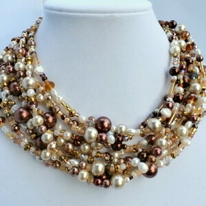 May include: A multi-strand statement necklace featuring an assortment of beads in shades of cream, champagne, bronze, and dark brown. The design incorporates round faux pearls, faceted crystal beads, and small glass seed beads in a layered arrangement.