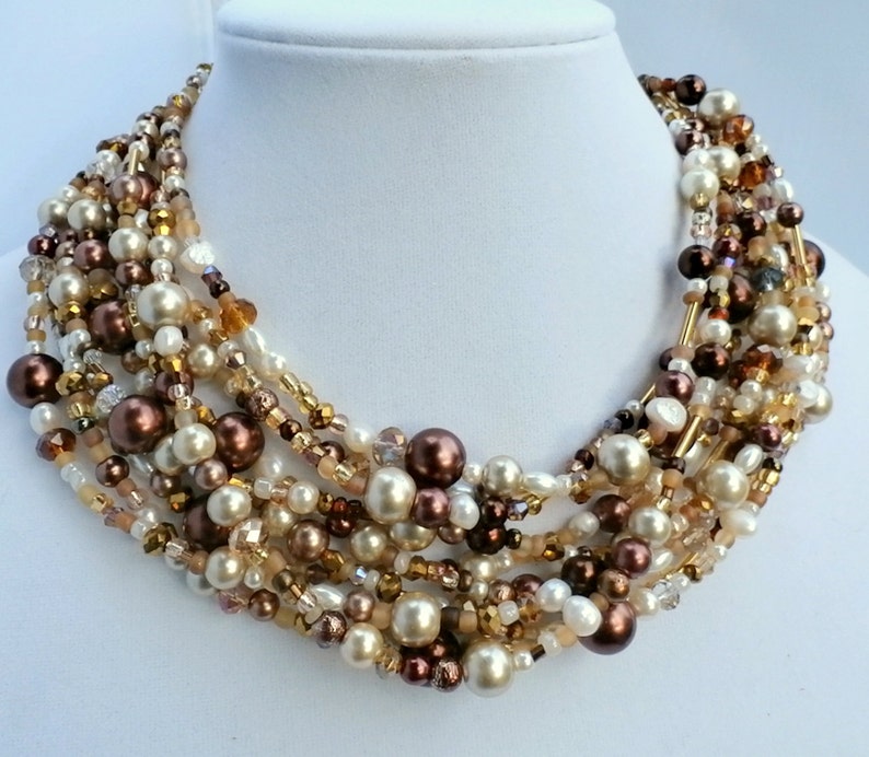 May include: A multi-strand statement necklace featuring an assortment of beads in shades of cream, champagne, bronze, and dark brown. The design incorporates round faux pearls, faceted crystal beads, and small glass seed beads in a layered arrangement.