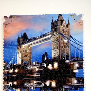 May include: A jigsaw puzzle featuring a colorful image of the Tower Bridge in London, England. The bridge is reflected in the water below, creating a symmetrical design. The puzzle pieces are visible in the image.