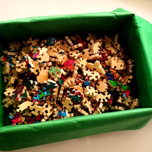 Peut inclure: Une bo&icirc;te verte remplie de pi&egrave;ces de puzzle en bois color&eacute;es. Les pi&egrave;ces sont de formes et de tailles diff&eacute;rentes et pr&eacute;sentent une vari&eacute;t&eacute; de motifs.