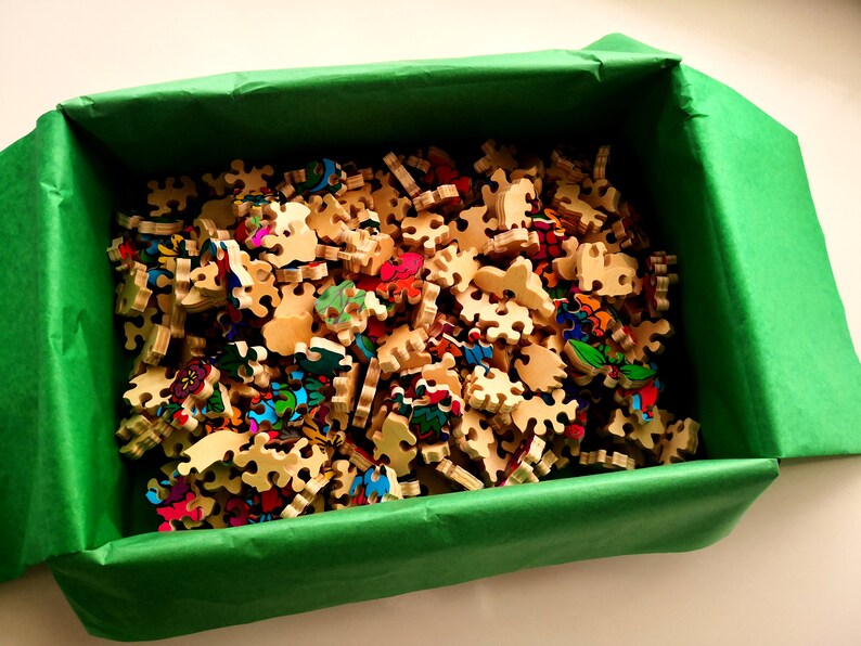Peut inclure: Une bo&icirc;te verte remplie de pi&egrave;ces de puzzle en bois color&eacute;es. Les pi&egrave;ces sont de formes et de tailles diff&eacute;rentes et pr&eacute;sentent une vari&eacute;t&eacute; de motifs.