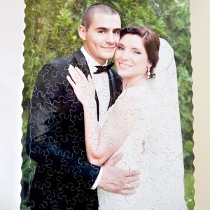 May include: A wedding photo puzzle featuring a couple in formal attire. The groom is wearing a black suit with a bow tie, and the bride is wearing a white dress with a veil. The puzzle is made of wood and has a scalloped edge.