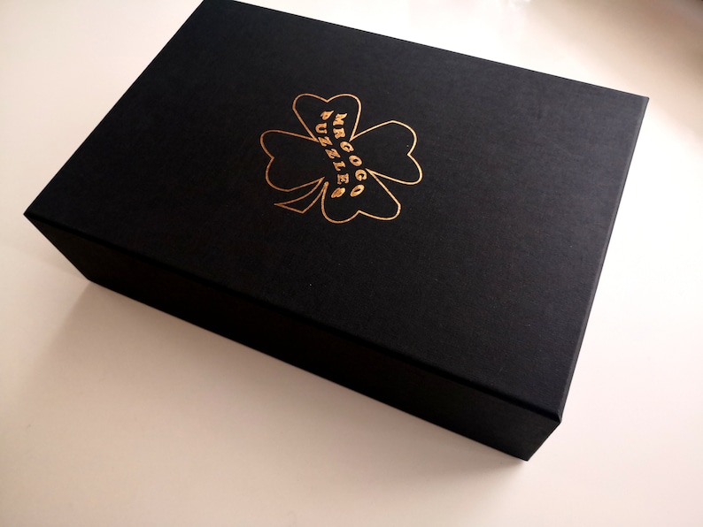 Peut inclure: Une bo&icirc;te noire avec un tr&egrave;fle &agrave; quatre feuilles en or et le texte "MRGOGG PUZZLE" sur le dessus.