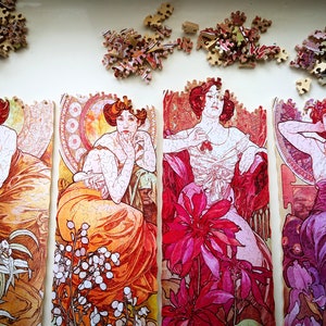 May include: Four jigsaw puzzle pieces featuring Art Nouveau style illustrations of women in elaborate dresses and floral designs. The pieces are in various shades of pink, red, orange, and purple.