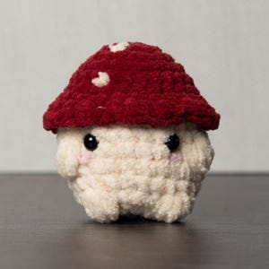 May include: A handmade crochet mushroom toy. The mushroom has a red cap with white spots and a cream-colored stem. The toy has small black eyes and a cute, friendly expression. It is a whimsical, soft toy.