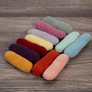 Könnte beinhalten: Eine Sammlung farbenfroher, zylindrischer Häkel-Haaraccessoires in verschiedenen Farbtönen, darunter Gelb, Orange, Rot, Lila, Rosa und Blau. Die Artikel sind auf einer Holzoberfläche angeordnet und zeigen ihre strukturierte, handgefertigte Optik.