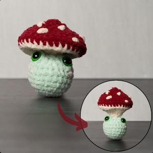 Könnte beinhalten: Ein handgehäkeltes Pilzspielzeug mit einem roten Hut mit weißen Punkten. Der Pilz hat einen hellgrünen Körper, grüne Augen und rosa Wangen. Das weiche, plüschige Spielzeug eignet sich für ein Kinderzimmer oder als dekoratives Element.