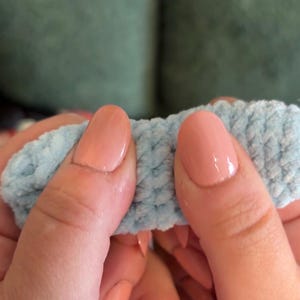 May include: A soft, light blue crocheted yarn piece with a textured, fuzzy appearance. The material is held between two thumbs, showcasing the dense, looped construction of the yarn. The light blue color is consistent throughout the visible section.