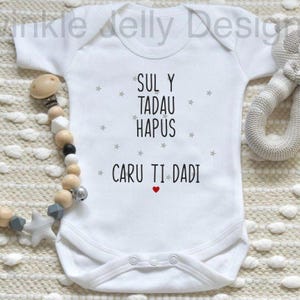 Puede incluir: Body de bebé blanco con el texto "SUL Y TADAU HAPUS CARU TI DADI" en negro, con pequeñas estrellas grises y un corazón rojo. A la izquierda hay un clip para chupete de madera y silicona. A la derecha hay un peluche.