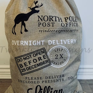 May include: A large beige burlap Christmas gift bag with a drawstring closure. The bag features a black silhouette of a reindeer and the words "North Pole Post Office" and "Overnight Delivery." Text on the bag includes "Do Not Open Before 25 December" and "Lillian."
