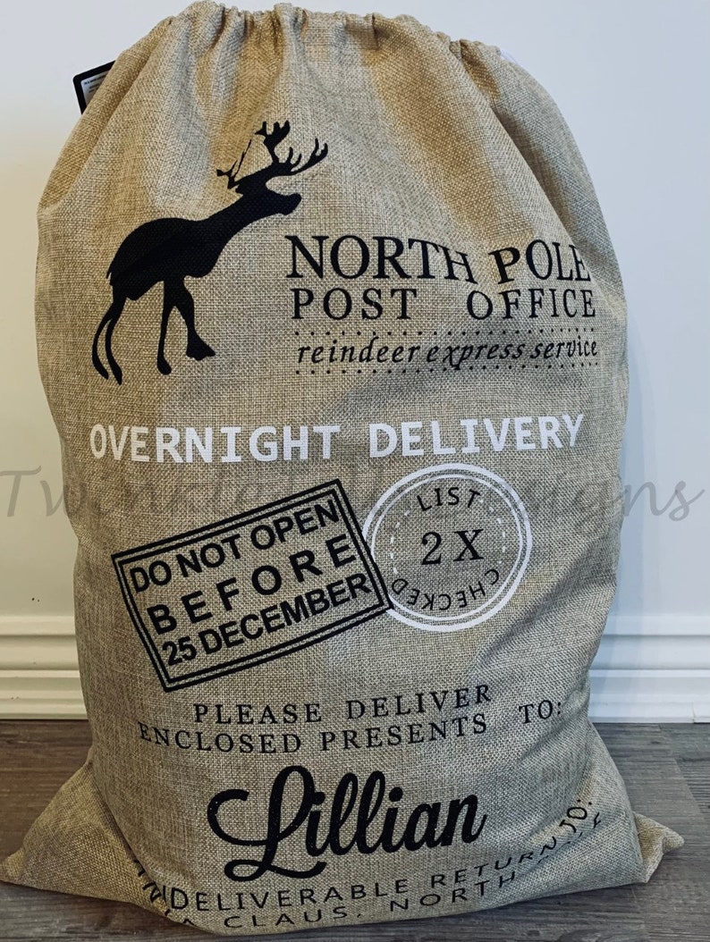 May include: A large beige burlap Christmas gift bag with a drawstring closure. The bag features a black silhouette of a reindeer and the words "North Pole Post Office" and "Overnight Delivery." Text on the bag includes "Do Not Open Before 25 December" and "Lillian."