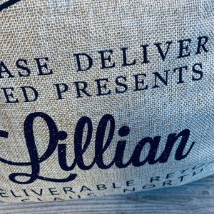 May include: Close-up of a burlap bag with the words "PLEASE DELIVER ED PRESENTS" in dark blue. The name "Lillian" is written in a cursive font with glitter. The bag has a rustic, textured appearance.