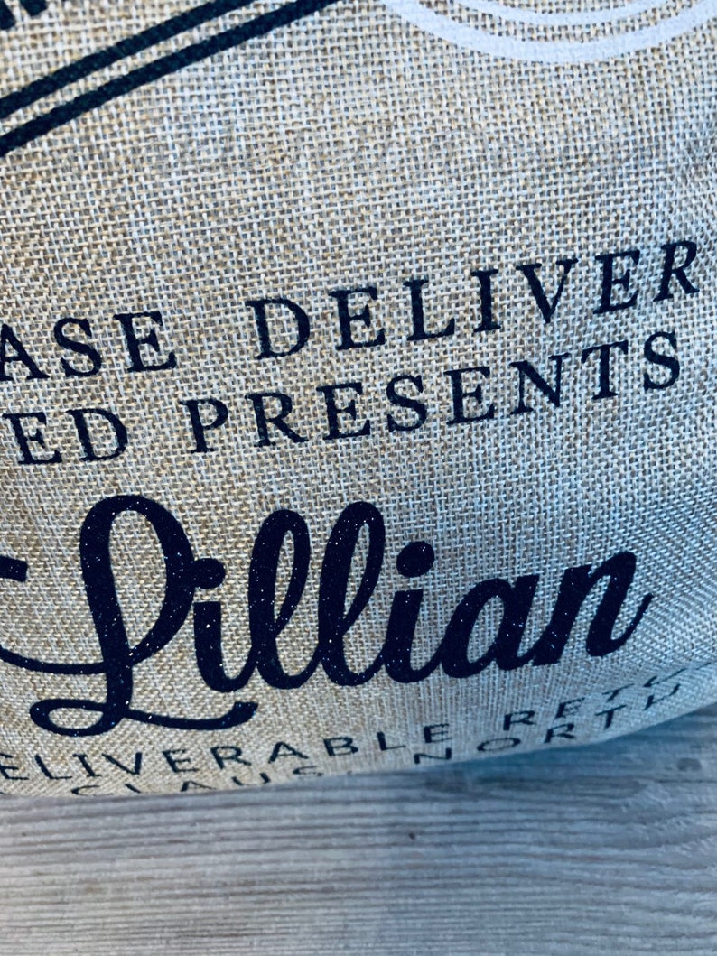 May include: Close-up of a burlap bag with the words "PLEASE DELIVER ED PRESENTS" in dark blue. The name "Lillian" is written in a cursive font with glitter. The bag has a rustic, textured appearance.