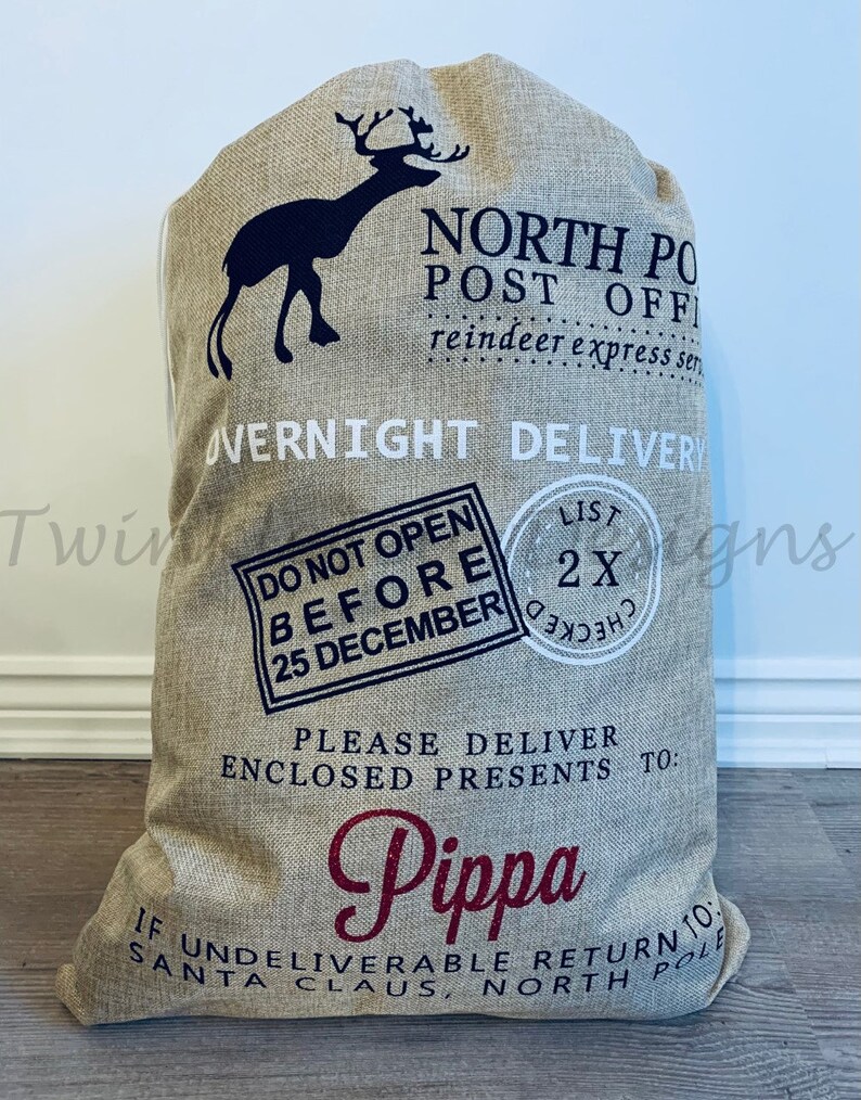 May include: A large beige burlap Santa sack with black and burgundy text and a reindeer silhouette. Text includes "North Pole Post Office," "Overnight Delivery," "Do Not Open Before 25 December," and "Pippa."