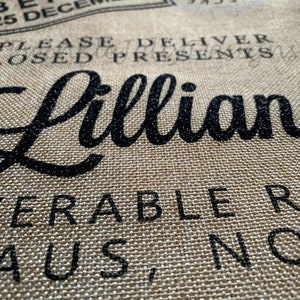 May include: Close-up of a burlap fabric with black text. The text includes the name "Lillian" in a cursive font, along with phrases like "Do Not Open Before 25 December" and "Please Deliver Closed Presents."